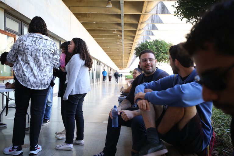 Estudantes conversam e aguardam sentados em um corredor de instituição de ensino.
